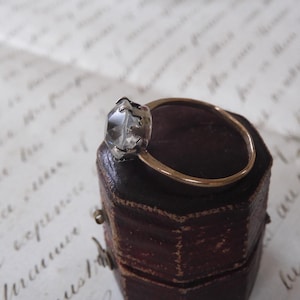 May include: A gold ring with a clear, faceted gemstone set in a silver-colored bezel. The ring is displayed on a small, dark brown, leather-like box, with a handwritten paper in the background.