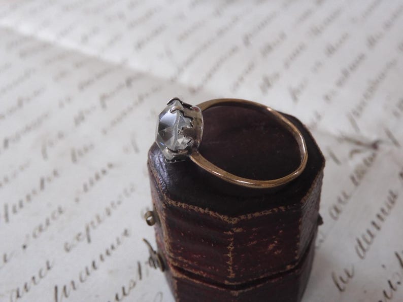May include: A gold ring with a clear, faceted gemstone set in a silver-colored bezel. The ring is displayed on a small, dark brown, leather-like box, with a handwritten paper in the background.