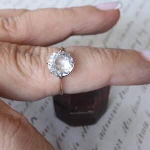 May include: A round, clear gemstone ring with a gold band. The large, faceted stone is the focal point, set in a classic prong setting. Another ring is visible in the background. The ring is displayed on a finger.