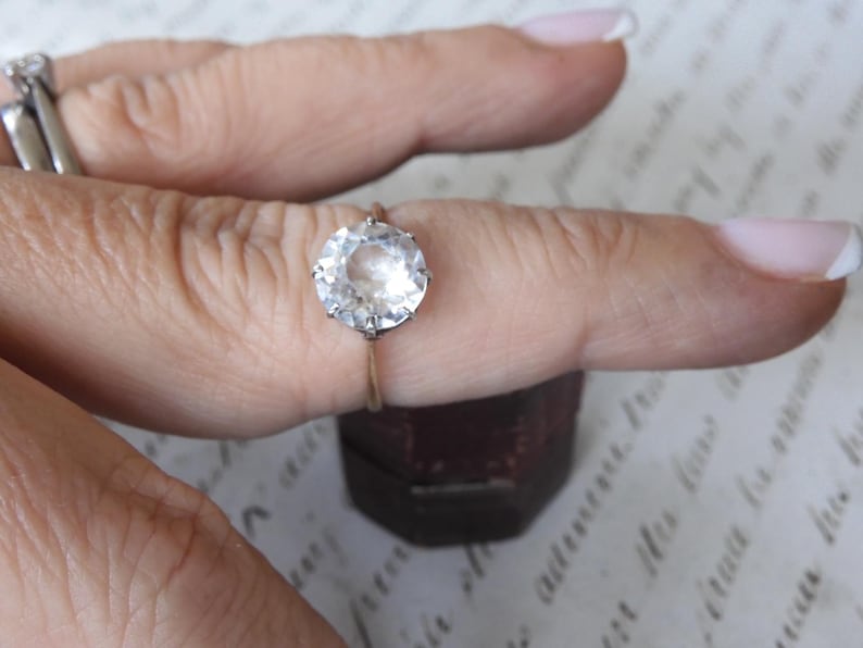 May include: A round, clear gemstone ring with a gold band. The large, faceted stone is the focal point, set in a classic prong setting. Another ring is visible in the background. The ring is displayed on a finger.