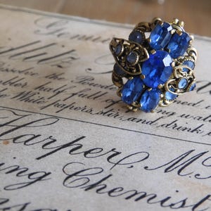 Peut inclure: Une bague vintage avec une grosse pierre bleue entourée de pierres bleues plus petites. La bague est en métal doré et présente un design détaillé.