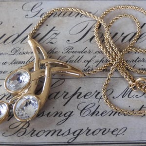 Könnte beinhalten: Goldfarbene Halskette mit einem Anhänger mit drei klaren, ovalen Kristallen. Der Anhänger hat ein abstraktes Design. Die Kette ist im zarten, gewebten Stil gehalten. Der Hintergrund ist ein Vintage-Papier mit verblasstem Text.
