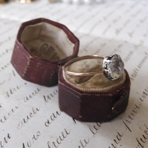 May include: An antique ring with a gold band and a silver-colored, flower-shaped setting, displayed in an open, burgundy-colored hexagonal ring box. The box sits on a vintage paper with handwritten script.