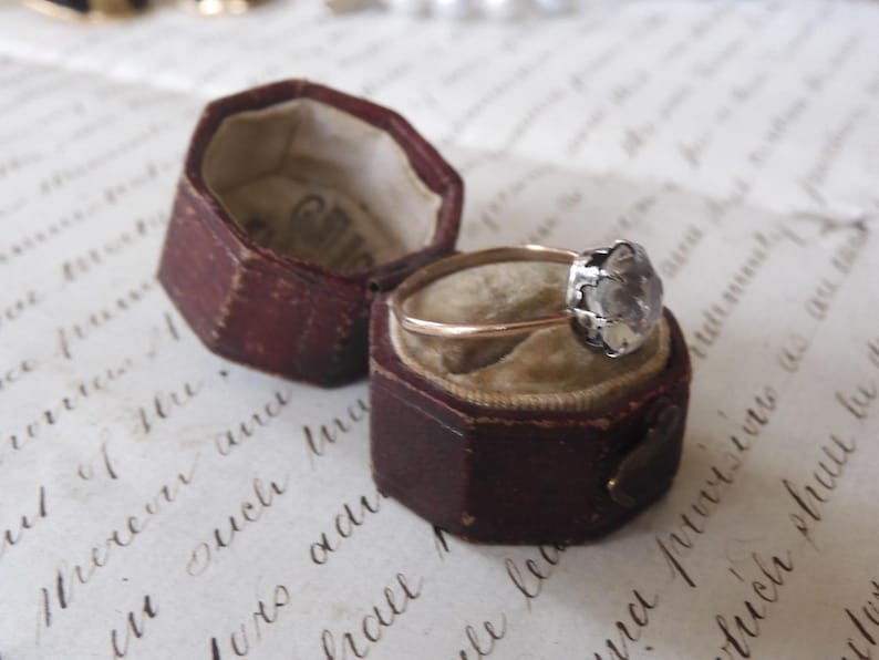 May include: An antique ring with a gold band and a silver-colored, flower-shaped setting, displayed in an open, burgundy-colored hexagonal ring box. The box sits on a vintage paper with handwritten script.