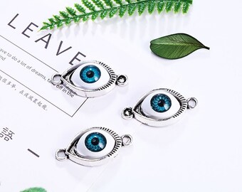 Puede incluir: Tres colgantes de plata con ojos azules. Cada colgante tiene forma de ojo con fondo blanco e iris azul. Los colgantes tienen un bucle a cada lado para sujetarlos a una cadena o pulsera.