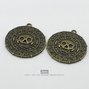 May include: Two antique gold-toned metal charms featuring a skull and crossbones design with intricate details. The charms are circular and have a raised edge.