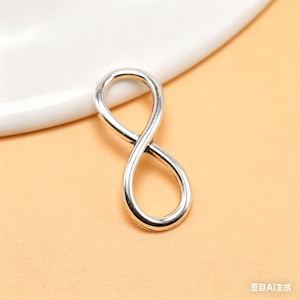 May include: A silver infinity symbol charm, set against a light orange backdrop. The charm's design is a continuous loop with a polished finish. A white, curved edge is visible in the background, possibly a plate or dish.