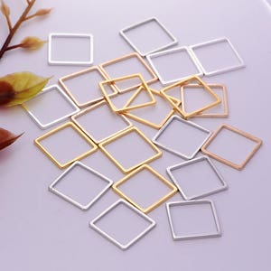 May include: A collection of square metal charms in silver, gold, and rose gold tones. The charms are open-framed and arranged on a light purple surface, suitable for jewelry making or crafting projects.