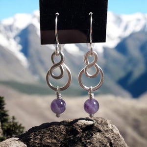 May include: Silver infinity symbol earrings with a purple amethyst gemstone dangle. The earrings are on a black background.