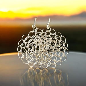 May include: A pair of silver earrings with a delicate, intricate design. The earrings are made of small, interconnected loops that create a circular shape. The earrings are hanging from simple silver hooks.