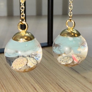 Puede incluir: Pendientes dorados con colgantes esféricos transparentes. Cada colgante contiene pequeñas conchas marinas y un diseño azul claro en forma de nube. Los pendientes cuelgan de finas cadenas doradas, con tema costero.