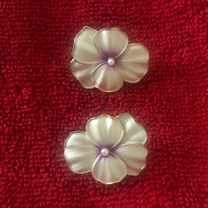 May include: A pair of flower-shaped pins featuring off-white petals with a subtle purple gradient toward the center. Each pin has a small, round, white bead at its core and a metallic gold-toned edge outlining the shape of the layered petals.