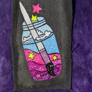 May include: A rectangular black fabric patch featuring a hand-painted design of a glass jar filled with blue, pink, and purple swirling liquid. A paintbrush is positioned diagonally inside the jar, surrounded by small neon yellow and pink stars.