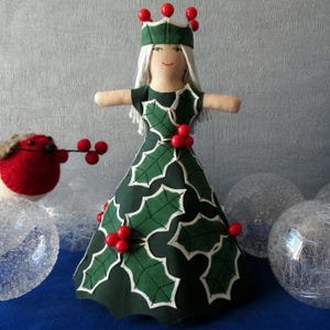 May include: A handmade Christmas tree topper doll in a green dress with white holly leaf details and red berry decorations. The doll has white hair, a green crown, and outstretched arms. A red bird with berries is also present.