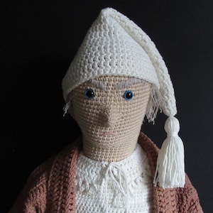 May include: A crocheted doll wearing a white crocheted sleeping cap with a tassel. The doll has a brown face with blue eyes and a white crocheted shirt and brown crocheted jacket.