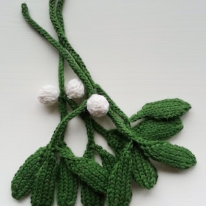 Könnte beinhalten: Ein handgestrickter Mistelzweig mit grünen Blättern und weißen Beeren. Der Zweig ist aus Garn gefertigt und perfekt zum Dekorieren für die Feiertage.