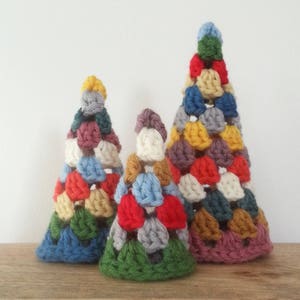 May include: Three crocheted Christmas trees in different sizes, made with colourful yarn. The largest tree is pink, brown, and yellow, the middle tree is green, red, and blue, and the smallest tree is blue, orange, and green.