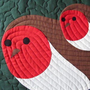 Puede incluir: Primer plano de un textil acolchado con dos petirrojos estilizados. Las aves tienen cuerpos rojos con ojos negros y picos marrones, sobre un fondo blanco y marrón. El fondo es una tela verde acolchada.