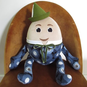 May include: A plush toy shaped like an egg with a green hat, blue and white patterned clothing, and a friendly face. The toy is sitting on a brown chair.