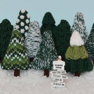 May include: A collection of knitted green and white Christmas trees with white snow at the base. A sign reads "North Pole" with distances to New York, Sydney, London, Los Angeles, and Tokyo.
