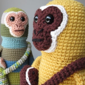 以下が含まれることがあります： 2つの編みぐるみのおさるさんの玩具。1つは黄色で、茶色の腕と白い顔をしています。もう1つは緑色、青色、茶色で、薄い茶色の顔をしています。