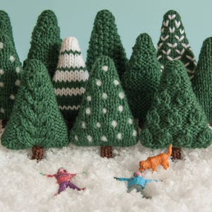 May include: A snowy scene with a group of knitted green Christmas trees, each with a different pattern. Two small figures are lying in the snow, and a small orange dog is standing near them.