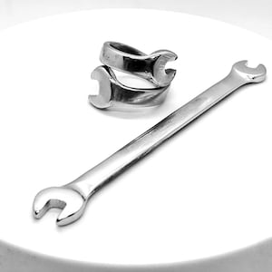 Può includere: Una chiave inglese argentata e due anelli a forma di chiave sono presentati su una superficie bianca. La chiave è lunga e sottile, con una testa curva ad ogni estremità. Gli anelli sono impilati, ognuno con un design a forma di chiave. Gli oggetti sono in metallo lucido.