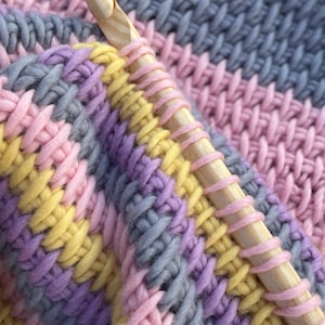 May include: Close-up of a crocheted blanket with a wooden crochet hook. The blanket is made with a variety of colors, including pink, yellow, purple, and gray.