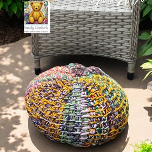 May include: A round, multi-colored crocheted pouf with yellow, purple, green, and white yarn. The pouf is on a concrete surface, with a gray wicker chair in the background. The image includes the text "Juey" and "Yarnstorm".