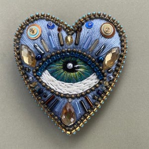 Puede incluir: Broche bordado en forma de coraz&oacute;n con dise&ntilde;o de ojo. El broche es principalmente azul claro, con un ojo detallado en tonos verdes y azules. Cuenta con cuentas, cristales y detalles met&aacute;licos, creando una pieza decorativa texturizada.