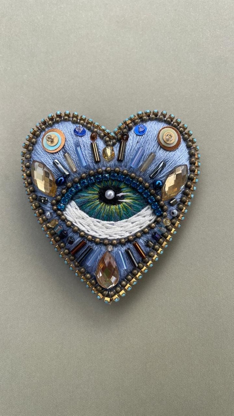 Puede incluir: Broche bordado en forma de coraz&oacute;n con dise&ntilde;o de ojo. El broche es principalmente azul claro, con un ojo detallado en tonos verdes y azules. Cuenta con cuentas, cristales y detalles met&aacute;licos, creando una pieza decorativa texturizada.