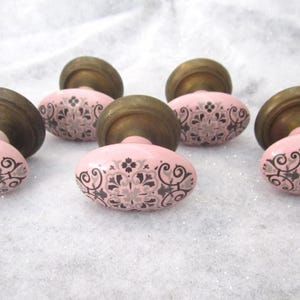 May include: A set of five oval-shaped cabinet knobs featuring a light pink ceramic surface with a dark gray and light gray floral pattern. Each knob is attached to a circular, antique-style brass base with a metallic finish and a rounded profile.