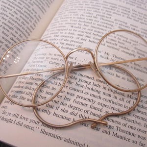 May include: Round gold-toned metal eyeglasses with thin frames and clear lenses resting on an open book with printed text. The glasses feature a classic bridge design and curved temple arms that are positioned directly on the paper pages.