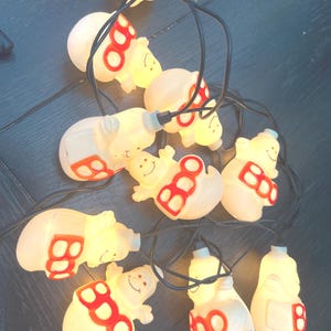 May include: A string of Halloween lights featuring white ghost-shaped bulbs with the word "BOO" in red. The lights are connected by a black wire and are lit up, creating a festive and spooky atmosphere.