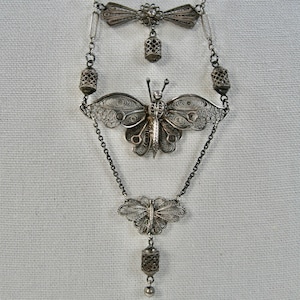 May include: A silver filigree necklace featuring three butterfly charms. The necklace has a delicate chain and a small bell charm at the bottom.