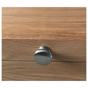 May include: A circular, brushed silver metal knob is attached to the front of a light brown wooden drawer. The wood features a visible horizontal grain pattern, and the knob has a smooth, reflective surface centered on the drawer front.