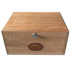 May include: A rectangular wooden box with a light brown grain, featuring box joint corners, a silver-coloured metal knob on the front lid, and an oval leather plaque centred on the front panel with the text Oscar Victor Baylias Treasure.