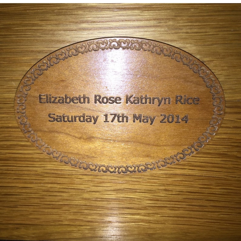 May include: An oval wooden plaque with a decorative border, featuring the engraved text Elizabeth Rose Kathryn Rice and the date Saturday 17th May 2014. The plaque is set against a light brown wood grain background with a smooth, polished finish.