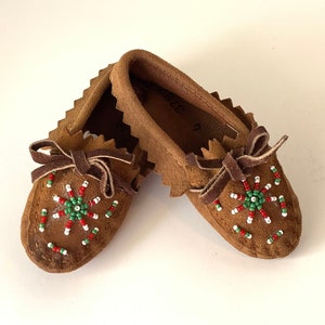 May include: A pair of brown leather moccasins with beaded designs. The moccasins have green, red, and white beads in a circular pattern.