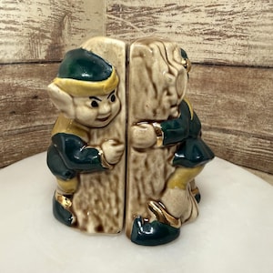 May include: A pair of ceramic bookends featuring two elf-like figures with pointed ears, wearing dark green and yellow outfits with gold accents. Each figure is sculpted to appear as if it is leaning against or climbing a textured, light brown tree trunk.