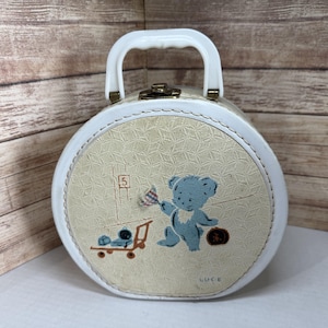 May include: Vintage round suitcase with a white handle and trim. The beige front features a blue teddy bear illustration with a flag and a toy wagon. The suitcase has a gold clasp and the word "GUCE" is printed on the bottom right.