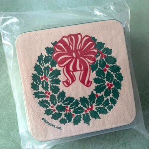 May include: A set of four beige coasters with a green holly wreath and a red bow design. The coasters are packaged in a clear plastic bag and have the text "CURRENT, INC." printed on the bottom.