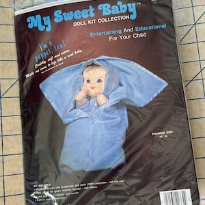 May include: A doll kit titled "My Sweet Baby" with a baby doll in a blue outfit. The kit includes a printed face, hair, and instructions. The finished size is 16 inches high.