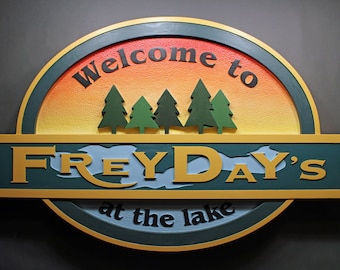 Może przedstawiać: Drewniany znak w zielono-złoto-niebieskiej kolorystyce. Na znaku widnieje napis "Welcome to FREYDAY'S at the lake" z zachodem słońca i sosnami w tle.