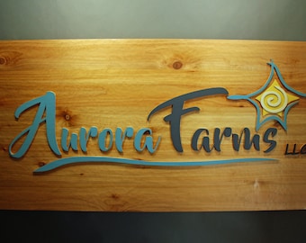 Może przedstawiać: Drewniany znak z tekstem "Aurora Farms LLC" w niebieskich i czarnych literach. Stylizowane słońce ze spiralą w środku znajduje się w prawym górnym rogu znaku.