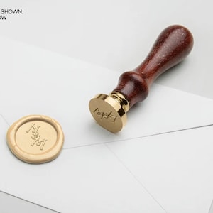 May include: A wooden handled wax seal stamp with a gold metal base. The stamp is imprinted with the letters "L & M" and is used to create a wax seal on a white envelope. The wax seal is a light yellow colour.