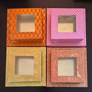 May include: Four decorative gift boxes in orange, pink, gold, and coral. Each box has a square window and a patterned design. The orange box has a paisley pattern, the pink box has white polka dots, the gold box has a floral design, and the coral box has a floral design.