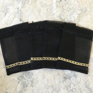 K&ouml;nnte beinhalten: Sechs schwarze Organza-Geschenkbeutel mit goldener Einfassung. Die Beutel sind in einer Reihe angeordnet und haben einen Kordelverschluss.