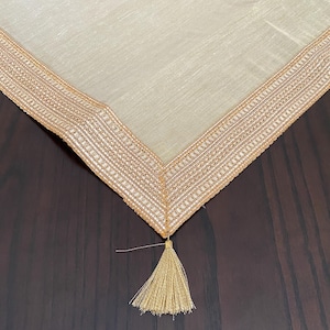 Könnte beinhalten: Ein hellbeiger Tischläufer mit strukturierter Stoffoberfläche und einer breiten, metallisch goldfarbenen Stickkante mit komplexen geometrischen Mustern. Die Ecke des Läufers ist mit einer einzelnen, passenden goldenen Quaste versehen.