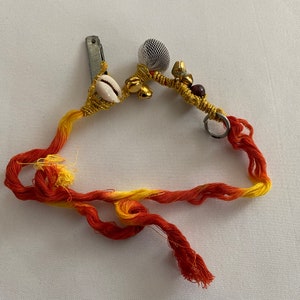 May include: A red and yellow string bracelet with a metal clasp, a white seashell, and gold-colored beads and charms.